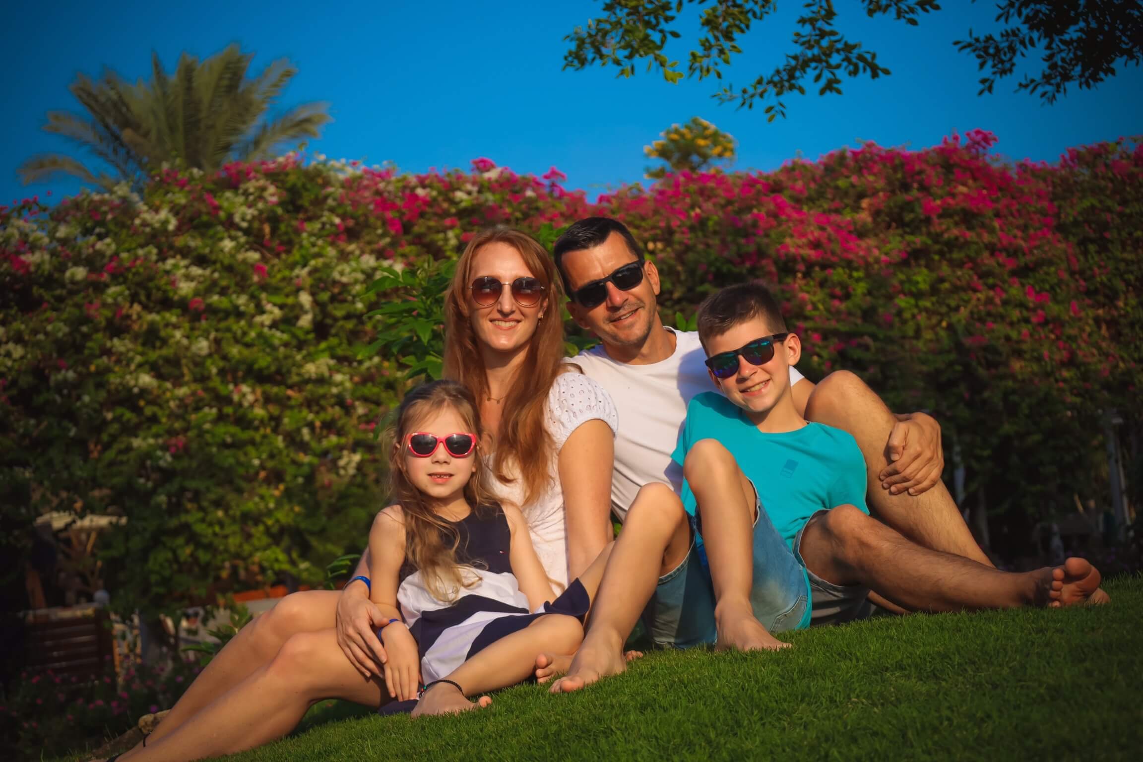 A family of four—father, mother, son, and daughter—are sitting on a green lawn and smiling at the camera. They are in a garden with a large bush of pink flowers behind them.